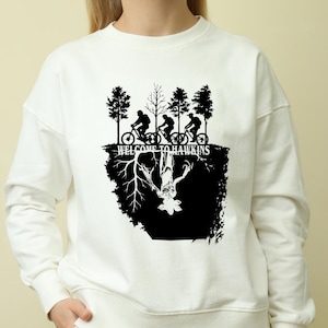 Op de afbeelding: Witte sweatshirt met een zwarte afbeelding van fietsers en bomen, de tekst "WELCOME TO HAWKINS" en een gespiegeld wezen. Het ontwerp is ge&iuml;nspireerd op een populaire televisieserie.
