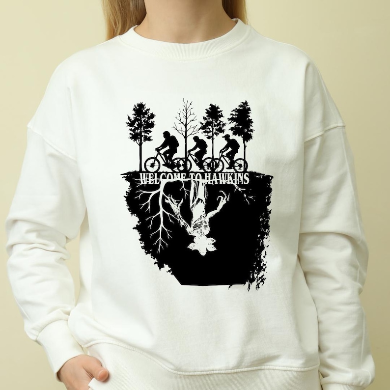 Op de afbeelding: Witte sweatshirt met een zwarte afbeelding van fietsers en bomen, de tekst "WELCOME TO HAWKINS" en een gespiegeld wezen. Het ontwerp is ge&iuml;nspireerd op een populaire televisieserie.