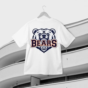 May include: White t-shirt featuring a bear's head graphic and the word "BEARS" in navy blue and orange. The shirt is suspended on a white hanger, set against a contemporary building. The bear graphic has an aggressive expression.