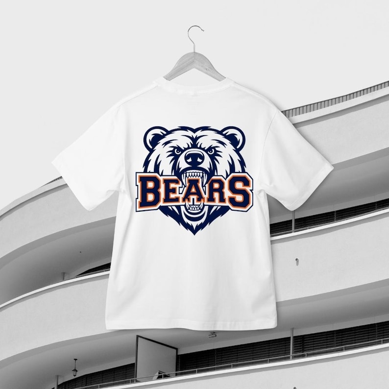 May include: White t-shirt featuring a bear's head graphic and the word "BEARS" in navy blue and orange. The shirt is suspended on a white hanger, set against a contemporary building. The bear graphic has an aggressive expression.