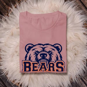 May include: A dusty rose t-shirt with a navy blue and orange bear graphic and the word "BEARS". The shirt is folded on a white fluffy surface, against a wooden backdrop. The design is bold and eye-catching.