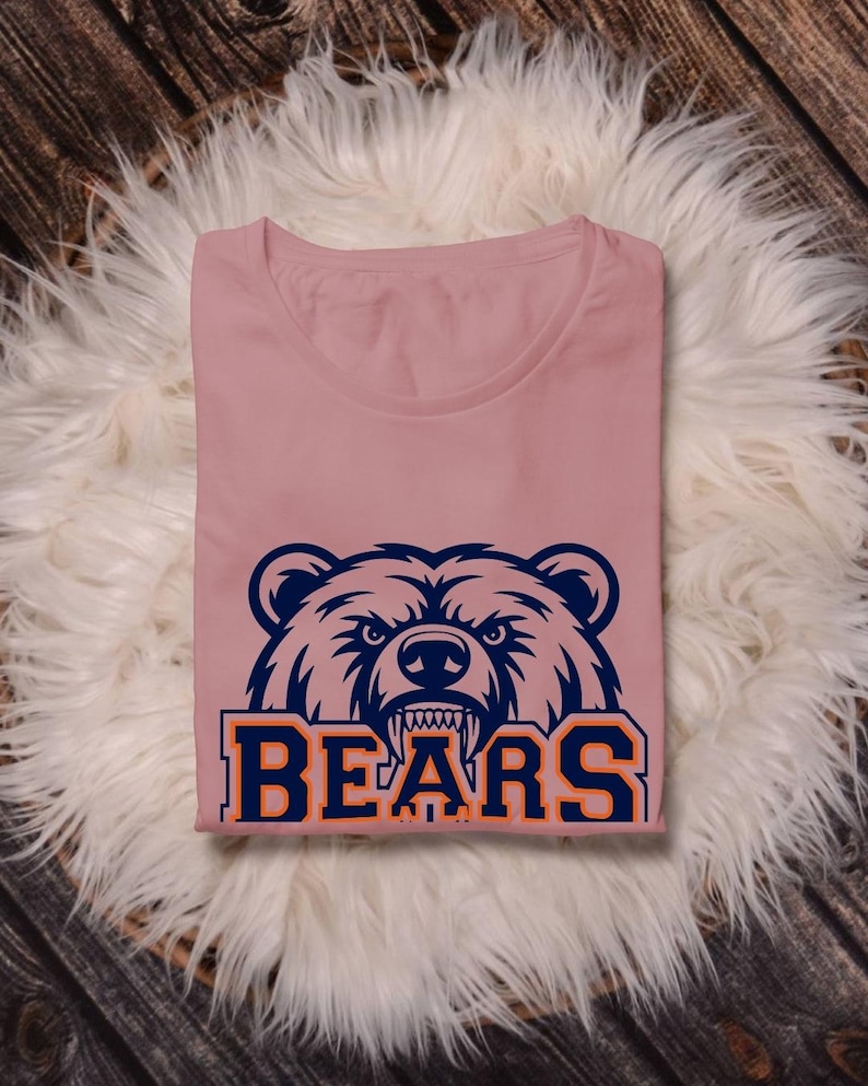 May include: A dusty rose t-shirt with a navy blue and orange bear graphic and the word "BEARS". The shirt is folded on a white fluffy surface, against a wooden backdrop. The design is bold and eye-catching.