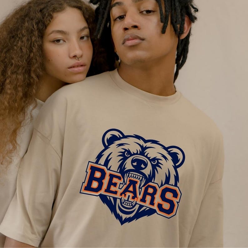 May include: Beige t-shirt featuring a navy blue and orange bear graphic and the word "BEARS". The graphic shows a detailed illustration of a bear's face with sharp teeth. The t-shirt is a casual style.