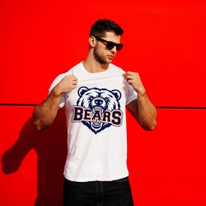 May include: White t-shirt with a navy and orange bear graphic and the word "BEARS". The design is set against a bright red background. The bear graphic has detailed fur and teeth. The t-shirt is being held up.