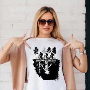 Op de afbeelding: Wit T-shirt met een zwarte afbeelding met fietsers, bomen en de tekst "WELCOME TO HAWKINS". Het ontwerp bevat een omgekeerde afbeelding van een wezen. Het T-shirt wordt door een persoon gedragen.