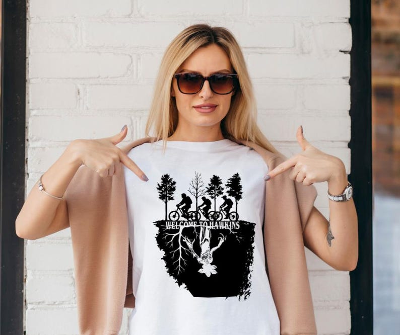Op de afbeelding: Wit T-shirt met een zwarte afbeelding met fietsers, bomen en de tekst "WELCOME TO HAWKINS". Het ontwerp bevat een omgekeerde afbeelding van een wezen. Het T-shirt wordt door een persoon gedragen.