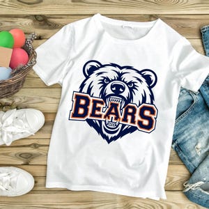 May include: White t-shirt with a navy and orange bear head graphic and the word "BEARS". The shirt is displayed with blue denim jeans. The image is set against a wooden background.