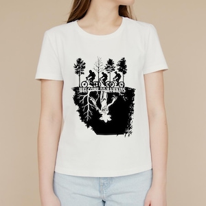 Op de afbeelding: Wit T-shirt met een zwarte afbeelding van fietsers en bomen boven een omgekeerd wezen en de tekst "WELCOME TO HAWKINS". Het ontwerp is in silhouetstijl.