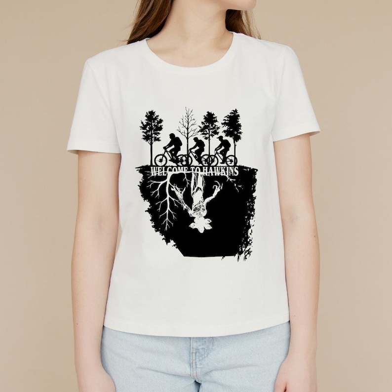 Op de afbeelding: Wit T-shirt met een zwarte afbeelding van fietsers en bomen boven een omgekeerd wezen en de tekst "WELCOME TO HAWKINS". Het ontwerp is in silhouetstijl.