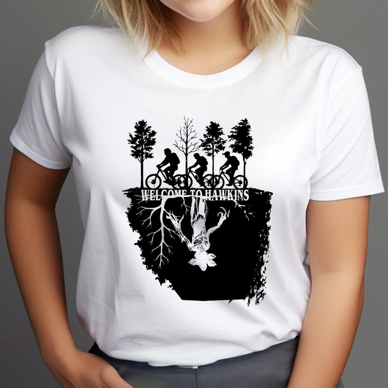 Op de afbeelding: Wit T-shirt met een zwart grafisch ontwerp uit de tv-serie Stranger Things. Het ontwerp bevat silhouetten van mensen op fietsen, bomen en de tekst "WELCOME TO HAWKINS". Hieronder een afbeelding van een monster.