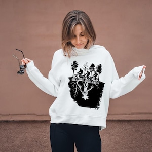 Op de afbeelding: Witte hoodie met een zwarte afbeelding met silhouetten van fietsers, bomen en de tekst "WELCOME TO HAWKINS". Het ontwerp bevat een omgekeerde figuur. De hoodie wordt door een persoon gedragen.
