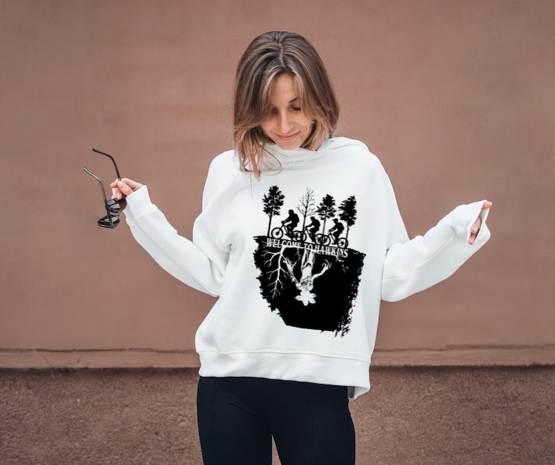 Op de afbeelding: Witte hoodie met een zwarte afbeelding met silhouetten van fietsers, bomen en de tekst "WELCOME TO HAWKINS". Het ontwerp bevat een omgekeerde figuur. De hoodie wordt door een persoon gedragen.