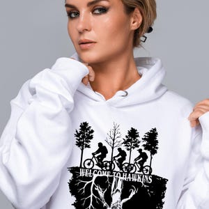 Op de afbeelding: Witte hoodie met een zwarte afbeelding van fietsers, bomen en de tekst "WELCOME TO HAWKINS". Het ontwerp staat op de voorkant. De hoodie heeft een capuchon met trekkoord en een kangoeroezak.