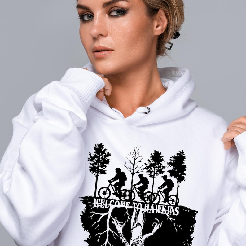 Op de afbeelding: Witte hoodie met een zwarte afbeelding van fietsers, bomen en de tekst "WELCOME TO HAWKINS". Het ontwerp staat op de voorkant. De hoodie heeft een capuchon met trekkoord en een kangoeroezak.