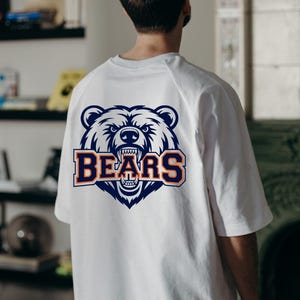 May include: White t-shirt featuring a navy blue and orange bear head graphic and the word "BEARS". The bear graphic has detailed fur and an open mouth. The shirt is a casual style.