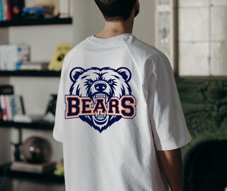 May include: White t-shirt featuring a navy blue and orange bear head graphic and the word "BEARS". The bear graphic has detailed fur and an open mouth. The shirt is a casual style.