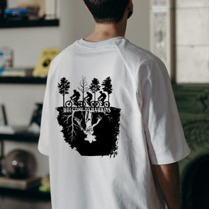 Op de afbeelding: Wit t-shirt met een zwarte afbeelding met silhouetten van fietsers, bomen en de tekst "WELCOME TO HAWKINS". Het ontwerp bevat een omgekeerde figuur en wortels, wat een unieke uitstraling cre&euml;ert.