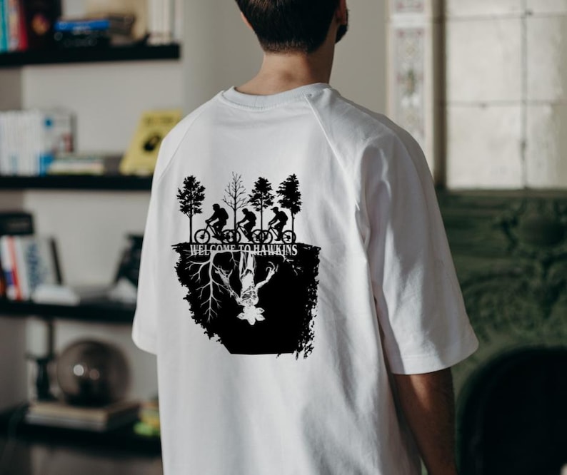 Op de afbeelding: Wit t-shirt met een zwarte afbeelding met silhouetten van fietsers, bomen en de tekst "WELCOME TO HAWKINS". Het ontwerp bevat een omgekeerde figuur en wortels, wat een unieke uitstraling cre&euml;ert.