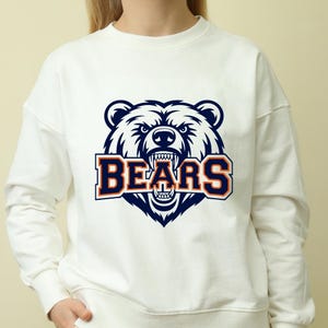 May include: White crew neck sweatshirt with a navy blue and orange bear head graphic and the word "BEARS". The bear is depicted with an angry expression and bared teeth. The sweatshirt is a casual style.