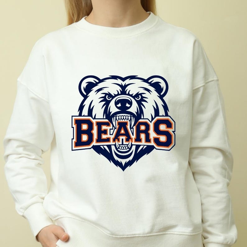 May include: White crew neck sweatshirt with a navy blue and orange bear head graphic and the word "BEARS". The bear is depicted with an angry expression and bared teeth. The sweatshirt is a casual style.