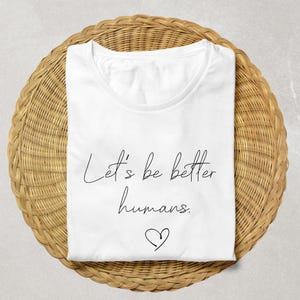 May include: A white t-shirt with the text "Let's be better humans" in a handwritten font, along with a heart symbol. The shirt is folded and placed on a woven, circular surface.