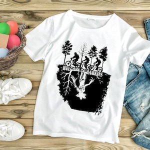 Op de afbeelding: Wit t-shirt met een zwart grafisch ontwerp. Het ontwerp bevat de tekst "WELCOME TO HAWKINS", silhouetten van fietsers en een monster. Het t-shirt is gemaakt van een zacht materiaal.