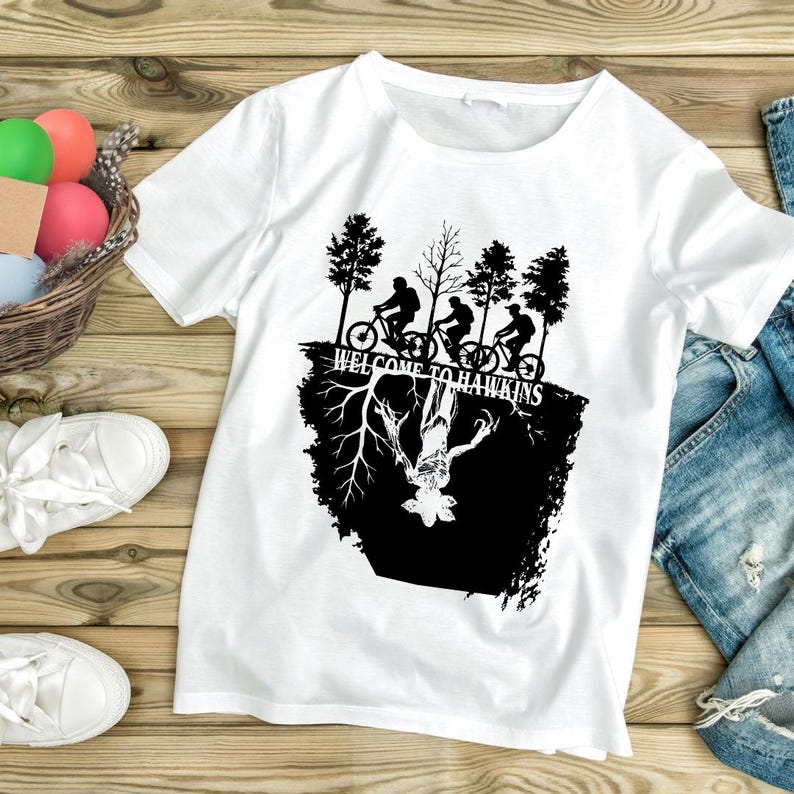 Op de afbeelding: Wit t-shirt met een zwart grafisch ontwerp. Het ontwerp bevat de tekst "WELCOME TO HAWKINS", silhouetten van fietsers en een monster. Het t-shirt is gemaakt van een zacht materiaal.
