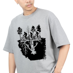 Op de afbeelding: Grijs crewneck T-shirt met een zwart grafisch ontwerp. Het ontwerp toont fietsers, bomen en de tekst "WELCOME TO HAWKINS". Daaronder een omgekeerde afbeelding van een monster, allemaal in zwart.