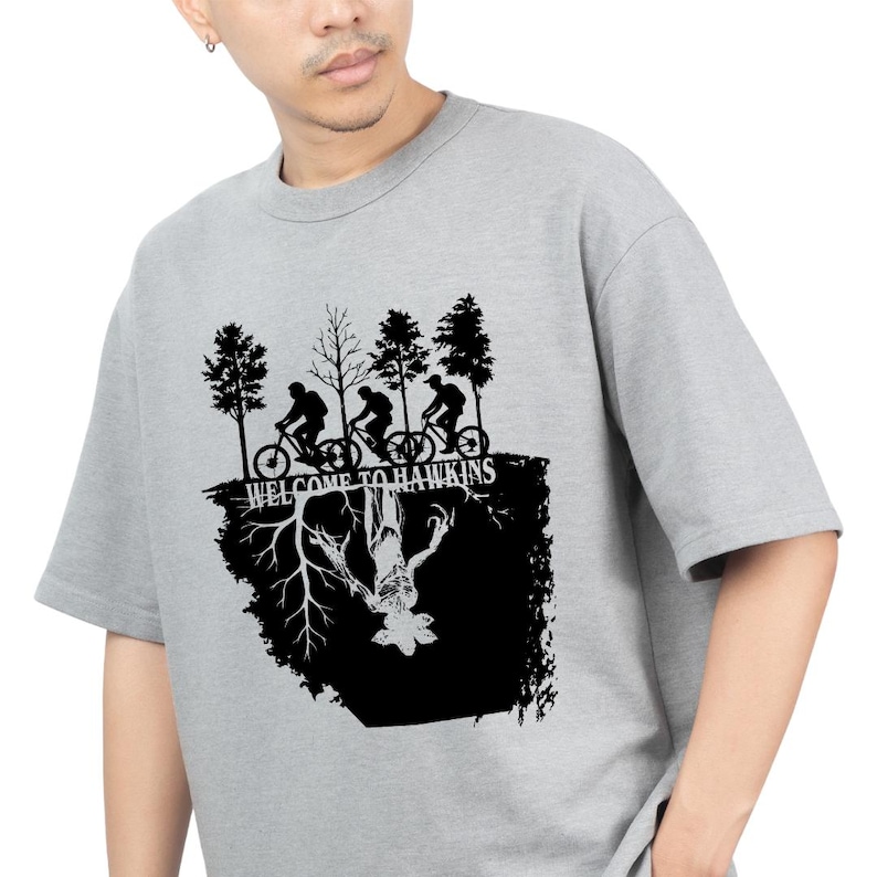 Op de afbeelding: Grijs crewneck T-shirt met een zwart grafisch ontwerp. Het ontwerp toont fietsers, bomen en de tekst "WELCOME TO HAWKINS". Daaronder een omgekeerde afbeelding van een monster, allemaal in zwart.