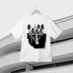 Op de afbeelding: Wit t-shirt met een zwarte print. Het ontwerp toont silhouetten van fietsers onder bomen, met de tekst "WELCOME TO HAWKINS". Daaronder een zwart omgekeerd beeld van een wezen en wortels.