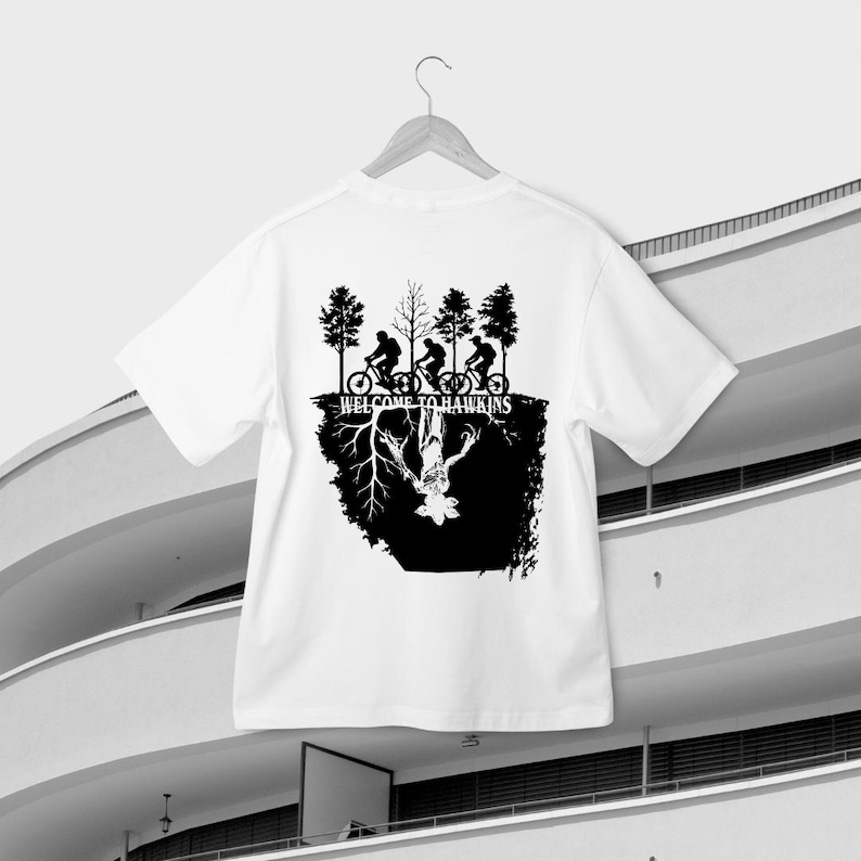 Op de afbeelding: Wit t-shirt met een zwarte print. Het ontwerp toont silhouetten van fietsers onder bomen, met de tekst "WELCOME TO HAWKINS". Daaronder een zwart omgekeerd beeld van een wezen en wortels.