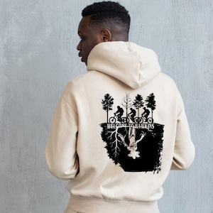 Op de afbeelding: Beige hoodie met een zwarte afbeelding op de rug. De afbeelding toont fietsers, bomen en de tekst "WELCOME TO HAWKINS". Het ontwerp bevat een omgekeerde figuur.