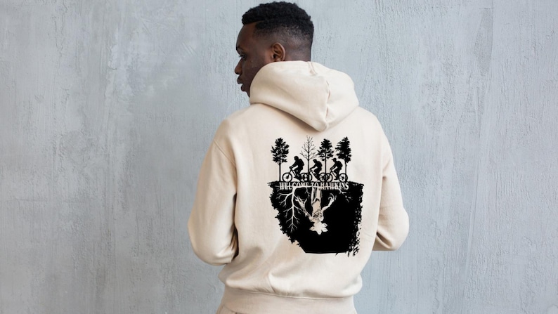 Op de afbeelding: Beige hoodie met een zwarte afbeelding op de rug. De afbeelding toont fietsers, bomen en de tekst "WELCOME TO HAWKINS". Het ontwerp bevat een omgekeerde figuur.