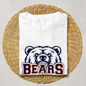 May include: White t-shirt with a navy and orange bear head graphic and the word "BEARS". The shirt is folded on a round, woven wicker surface. The design is a bold, eye-catching statement.