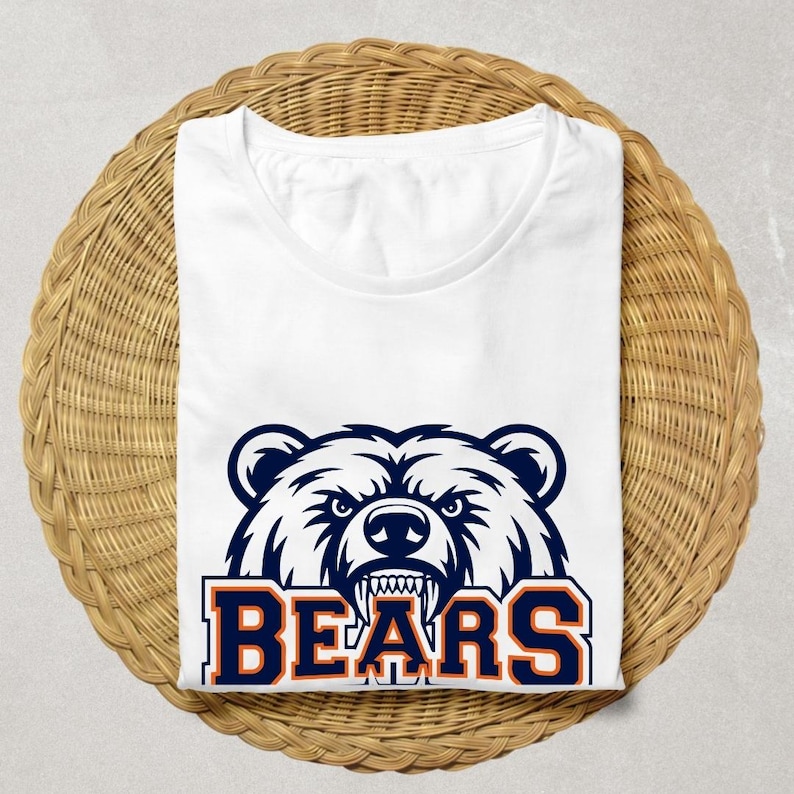May include: White t-shirt with a navy and orange bear head graphic and the word "BEARS". The shirt is folded on a round, woven wicker surface. The design is a bold, eye-catching statement.