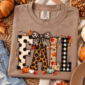 May include: A tan t-shirt featuring the word FALL in large letters. The F has black polka dots, the A has a leopard print with a bow, the L has autumn leaves, and the second L has a black and white buffalo check pattern with pumpkins.