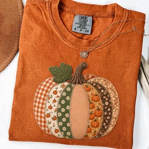May include: A burnt orange short-sleeve t-shirt featuring a large pumpkin applique on the front. The pumpkin is constructed from various fabric patches including gingham, floral, and herringbone patterns. The shirt has a Comfort Colors label.