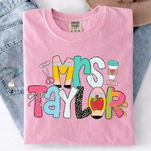 May include: Pink Comfort Colors t-shirt with the name "Mrs. Taylor" in colorful, whimsical letters. The design includes school-themed elements like pencils, scissors, an apple, and a coffee cup. The shirt is folded on a pair of blue jeans.