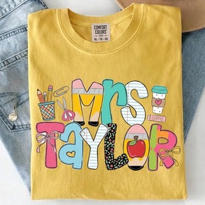 May include: Yellow Comfort Colors t-shirt with the name "Mrs. Taylor" in colorful, playful lettering. The design includes school-themed graphics like pencils, scissors, and a coffee cup. The shirt is folded on a pair of blue jeans.