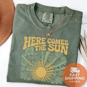 May include: An olive green short-sleeve t-shirt with a crew neckline. The shirt features a graphic print with the yellow text "HERE COMES THE SUN" above a stylized sunburst design surrounded by green floral illustrations on the front chest.