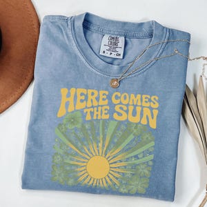 May include: A blue short-sleeve t-shirt with a graphic print featuring the yellow text "HERE COMES THE SUN" above a yellow sun and green floral design. The shirt has a Comfort Colors brand label at the inner neckline and a crew neck collar.