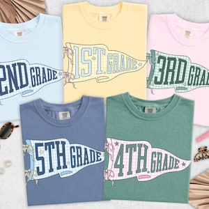 May include: Five crew neck t-shirts in light blue, yellow, pink, dark blue, and green. Each shirt features a pennant graphic with text reading 1st Grade, 2nd Grade, 3rd Grade, 4th Grade, and 5th Grade, decorated with small stars and ribbon bows.