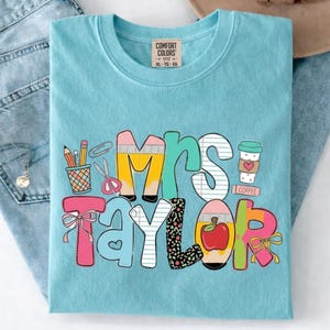 May include: A light blue t-shirt with a graphic print featuring the text Mrs. Taylor. The design includes colorful letters decorated with patterns like pencils, notebook paper, apples, and coffee cups, accented with pink bows and scissors.
