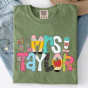 May include: An olive green t-shirt with a graphic print that reads Mrs. Taylor. The letters are decorated with school-themed illustrations including pencils, a cup, scissors, a red apple, and bows. The shirt has a Comfort Colors brand label at the neck.