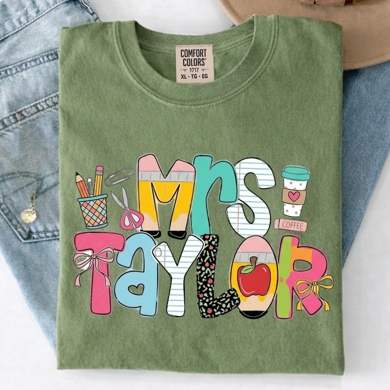 May include: An olive green t-shirt with a graphic print that reads Mrs. Taylor. The letters are decorated with school-themed illustrations including pencils, a cup, scissors, a red apple, and bows. The shirt has a Comfort Colors brand label at the neck.