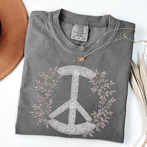 May include: A gray Comfort Colors t-shirt featuring a peace sign design with a floral wreath. The peace sign is filled with a glittery material. A silver necklace with a round pendant is draped across the shirt. A brown hat is visible in the upper left corner.