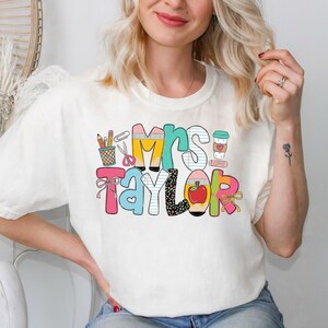 May include: A cream-colored short-sleeve t-shirt featuring a colorful graphic print that reads "Mrs. Taylor" in large, stylized letters. The design incorporates school-themed illustrations including a pencil cup, scissors, and an apple.
