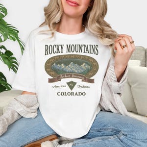 Pu&ograve; includere: T-shirt bianca con un design grafico. Il testo "Rocky Mountains" &egrave; sopra un'illustrazione ovale di montagne e alberi. Sotto, uno striscione con le parole "Outdoor Discovery". La maglietta include anche le parole "American Tradition" e "Colorado".