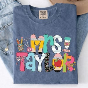 May include: A blue t-shirt featuring a graphic print with the text "Mrs. Taylor" in colorful, patterned letters. The design includes illustrations of a pencil holder, scissors, a coffee cup, and an apple, accented with pink and yellow bows.