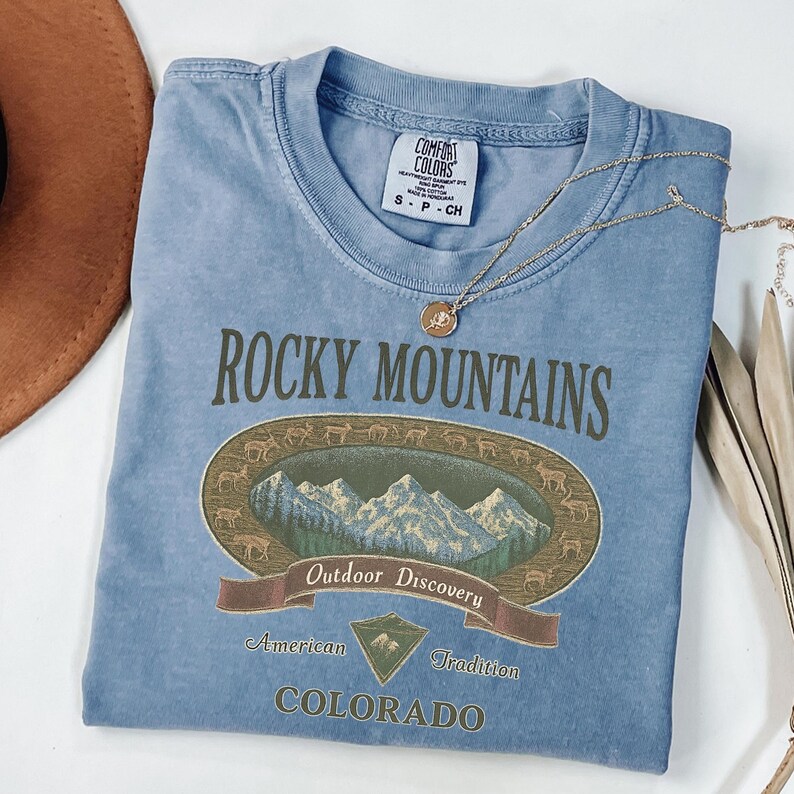 Pu&ograve; includere: Una maglietta azzurra con la scritta "Rocky Mountains" e una grafica di montagne e cervi. La maglietta riporta anche le parole "Outdoor Discovery", "American Tradition" e "Colorado". Una collana d'oro &egrave; drappeggiata sulla maglietta.